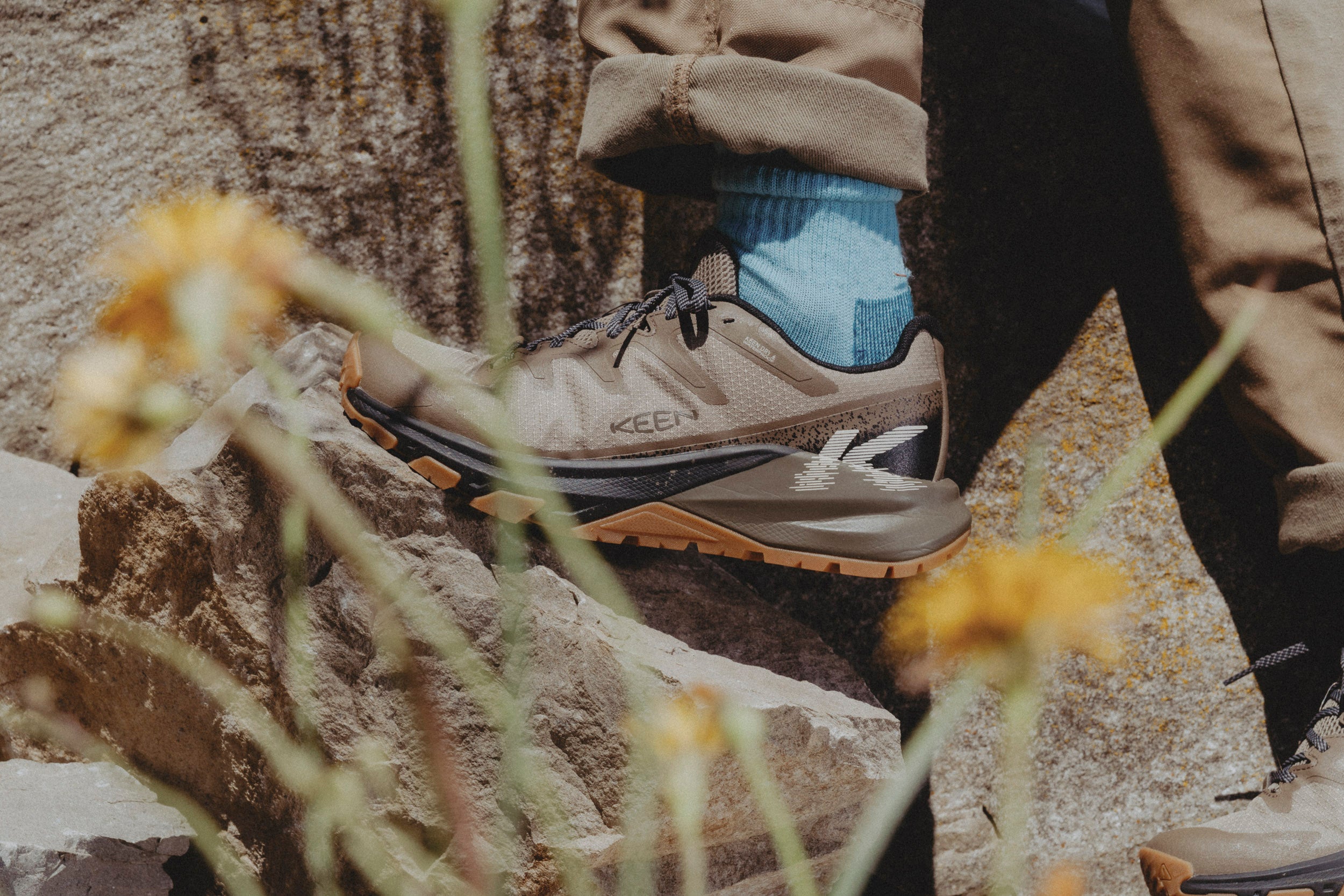 A close-up of a person hiking in rocky terrain, wearing a beige Keen targhee apex hiking shoe with a blue sock and brown pants.