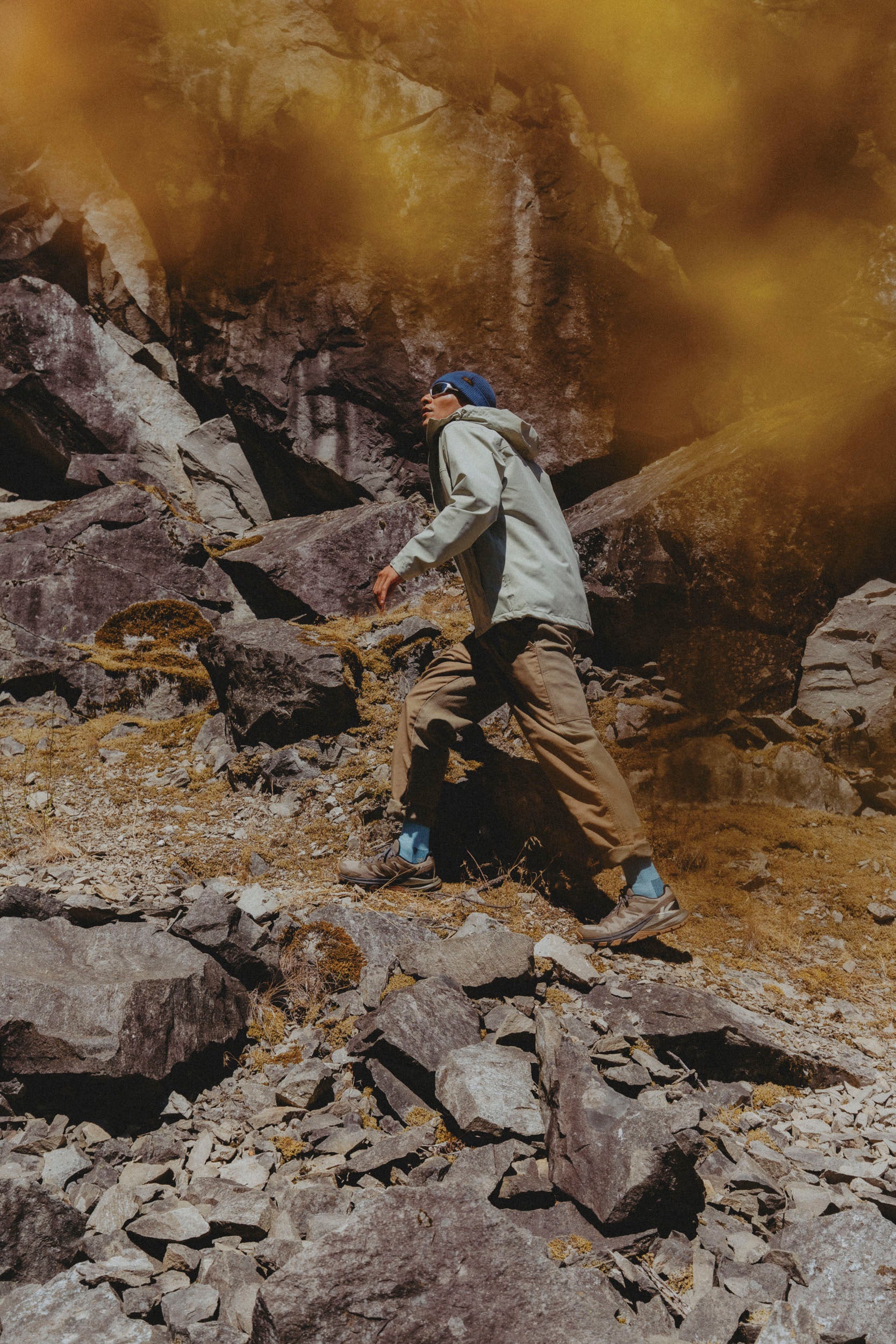 A person hiking through a rocky, dusty outdoor terrain under a warm golden sky, wearing a light gray hooded jacket, and keen targhee apex brown hiking shoes 