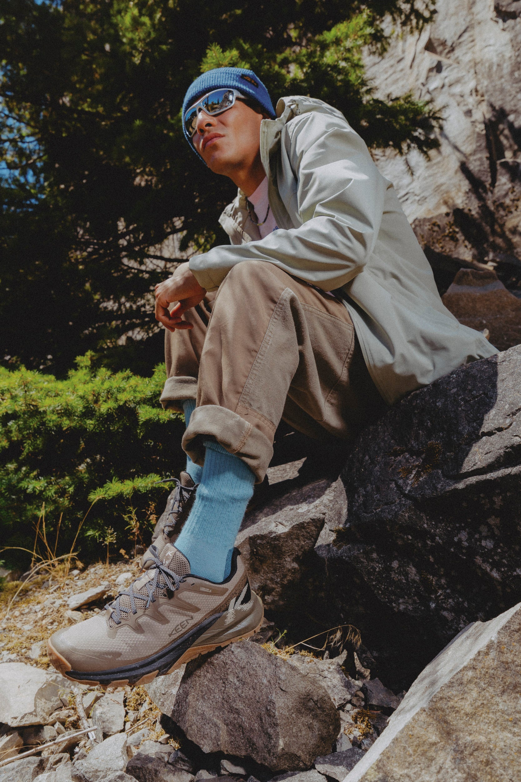 A man wearing a light beige outdoor jacket, brown cargo pants, light blue socks, and keen brown targhee apex hiking shoes sat on a large rock 