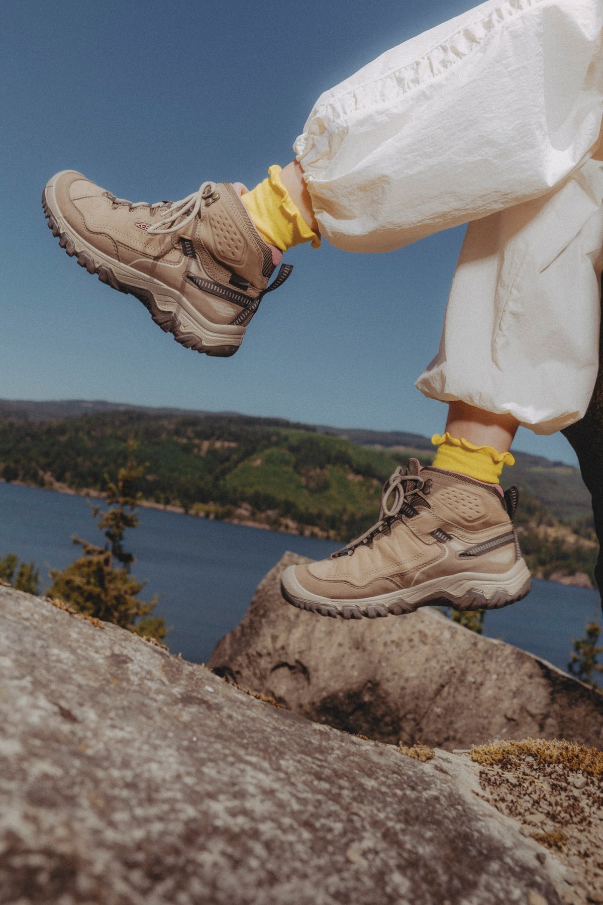 A person sitting on a rocky overlook with a lake and forest in the background, wearing keen targhee