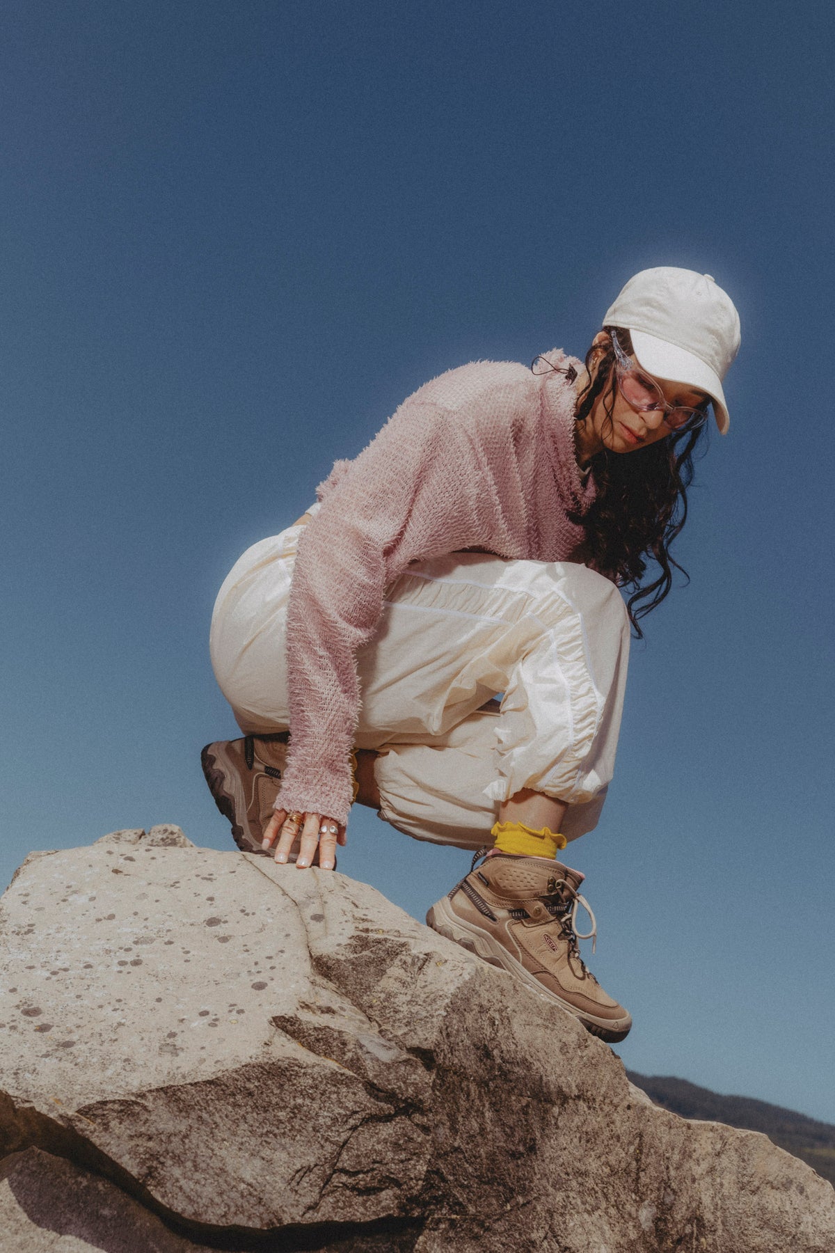 A person crouching on a large rock outdoors under a clear blue sky, wearing a white cap, a light