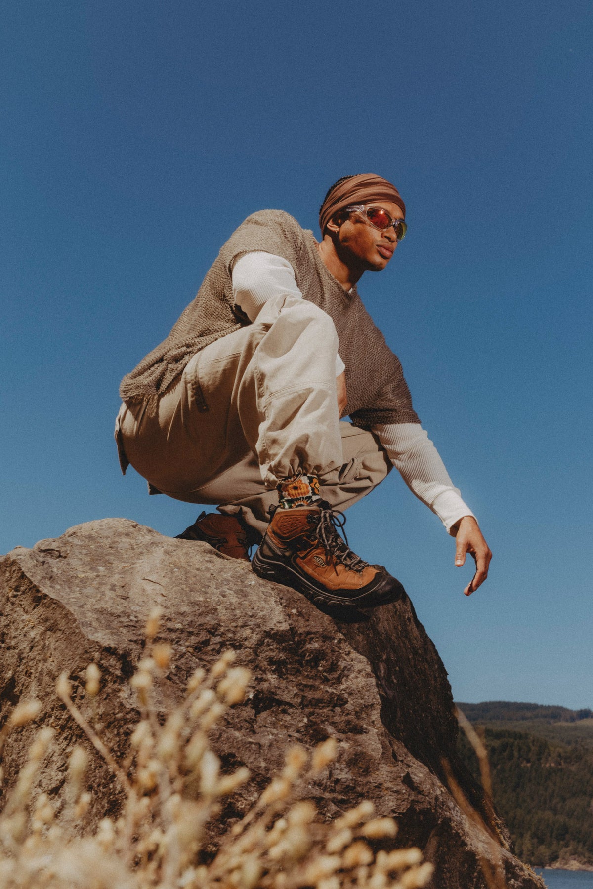 A man crouches on a large rock outdoors under a clear blue sky, wearing a brown knit sweater vest