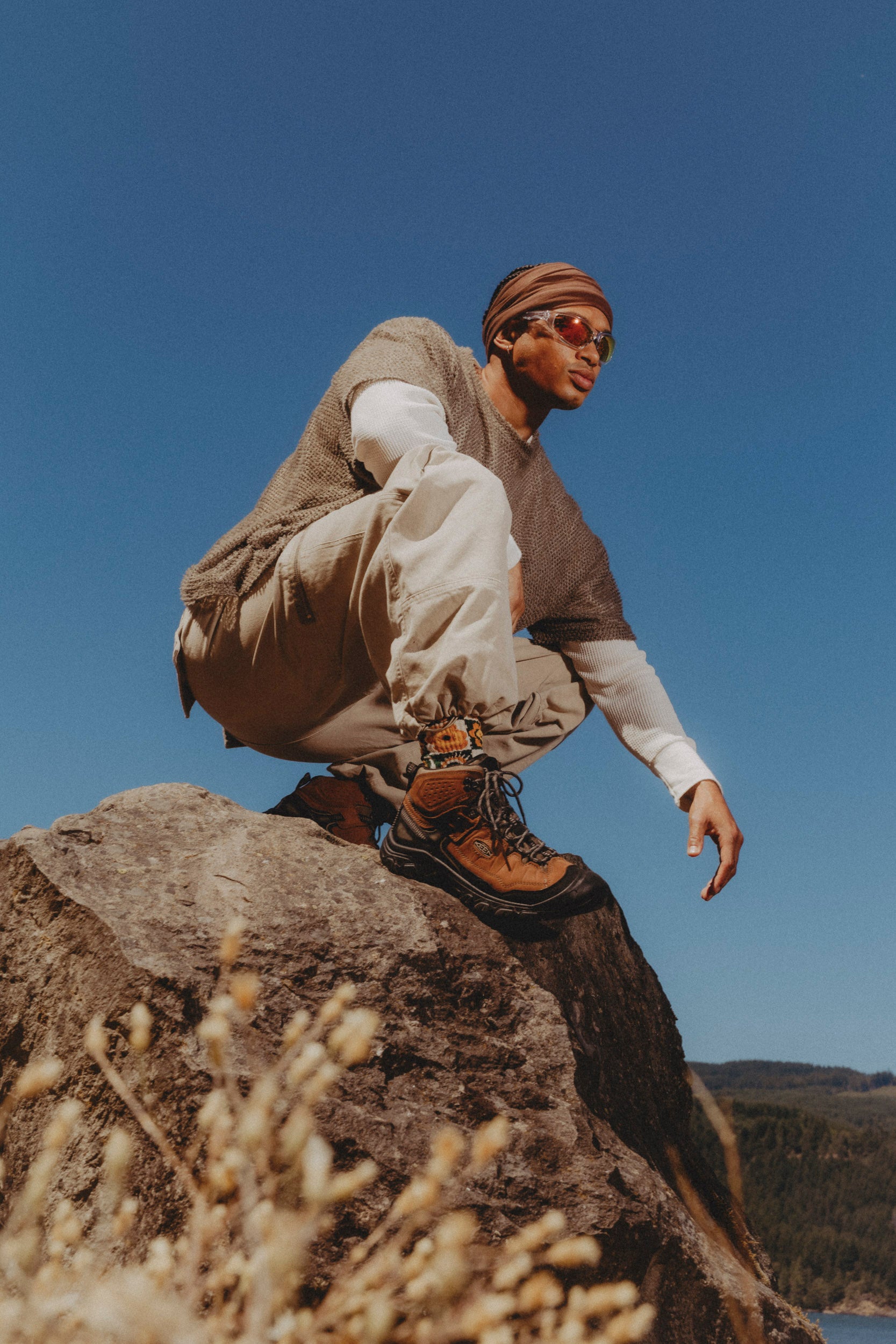 A man crouches on a large rock outdoors under a clear blue sky, wearing a brown knit sweater vest over a white long-sleeve shirt, beige pants, brown keen targhe IV hiking boots, and a brown beanie.