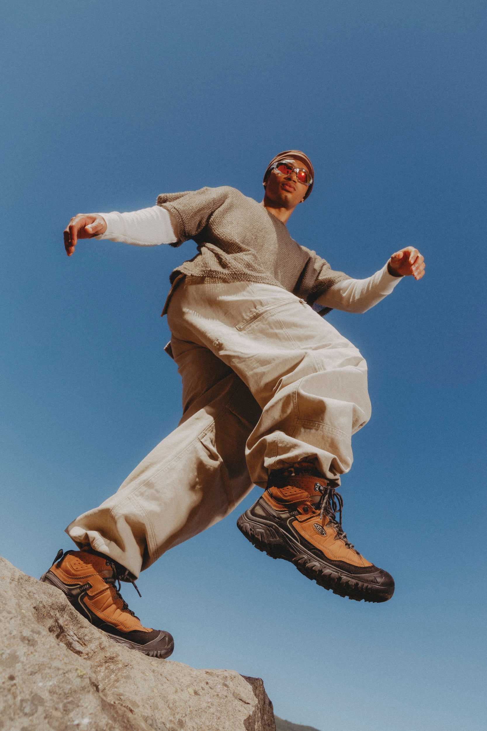 A person outdoors on a rock against a clear blue sky, wearing loose beige cargo-style clothing, a taupe long-sleeve top, brown keen targhee IV hiking boots, and sunglasses.