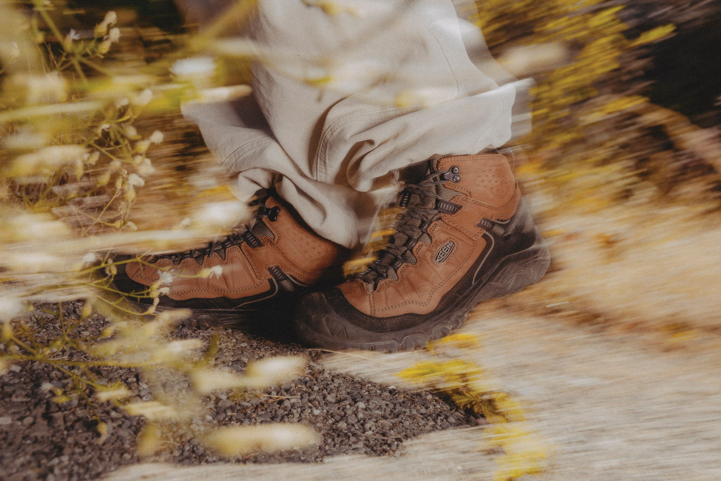A close-up action shot of a person wearing keen brown targhee IV hiking boots and light-colored pants, captured with a motion blur effect over yellow wildflowers.
