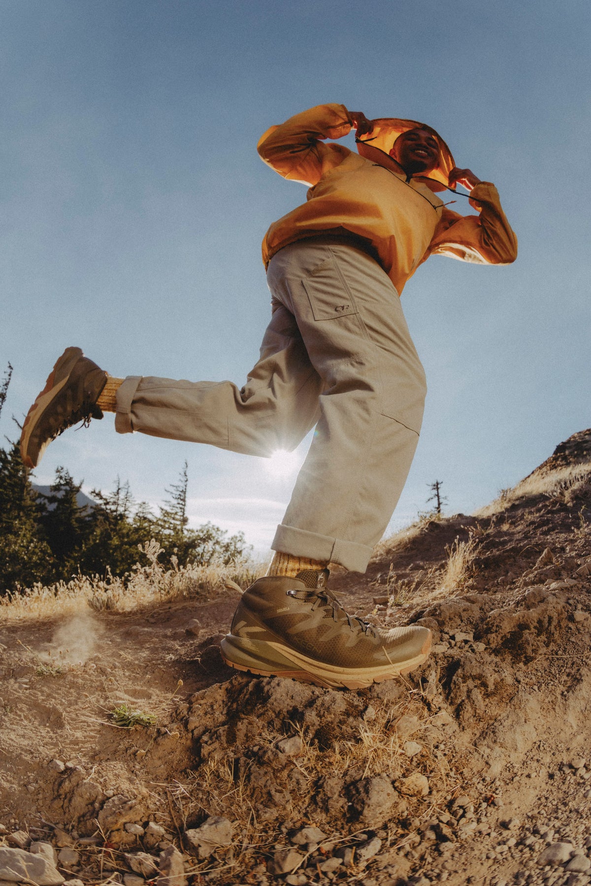 A person hiking on a rocky outdoor trail, wearing an orange long-sleeve top, light beige cargo