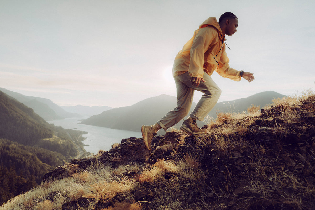A man in a yellow hoodie and beige pants hiking up a grassy mountain ridge with a scenic lake