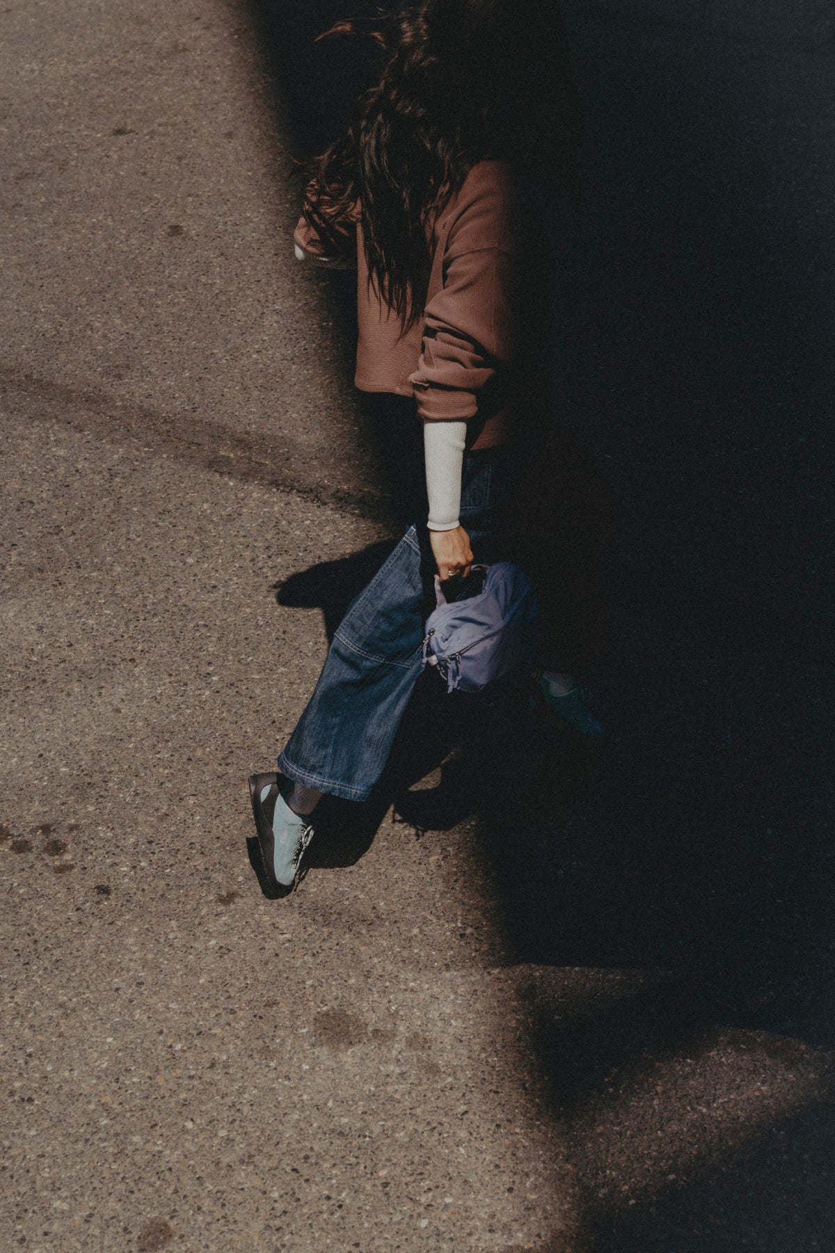 A high-angle shot of a person walking on a sunlit asphalt street, wearing a brown jacket, blue