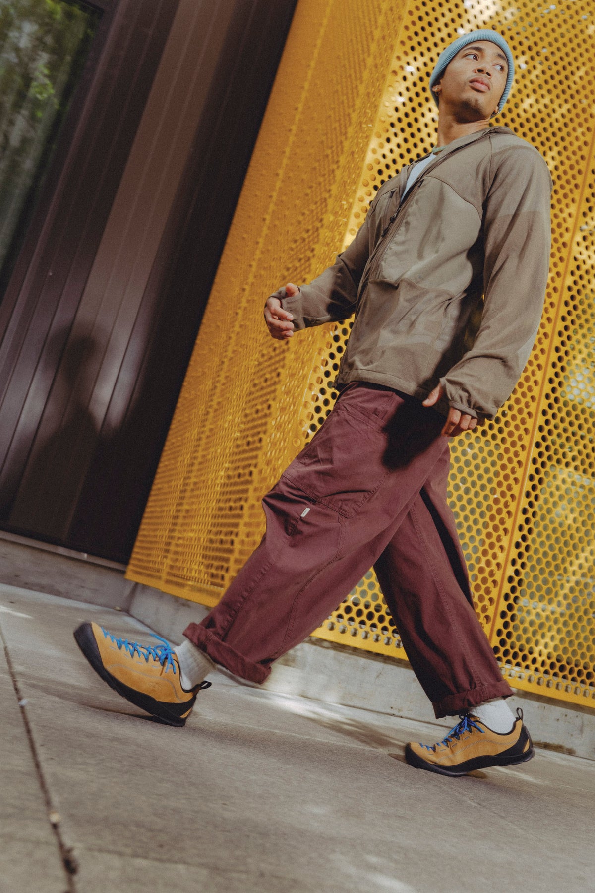 A man walking outdoors wearing a beige utility jacket, maroon cargo pants, and a brown jasper suede