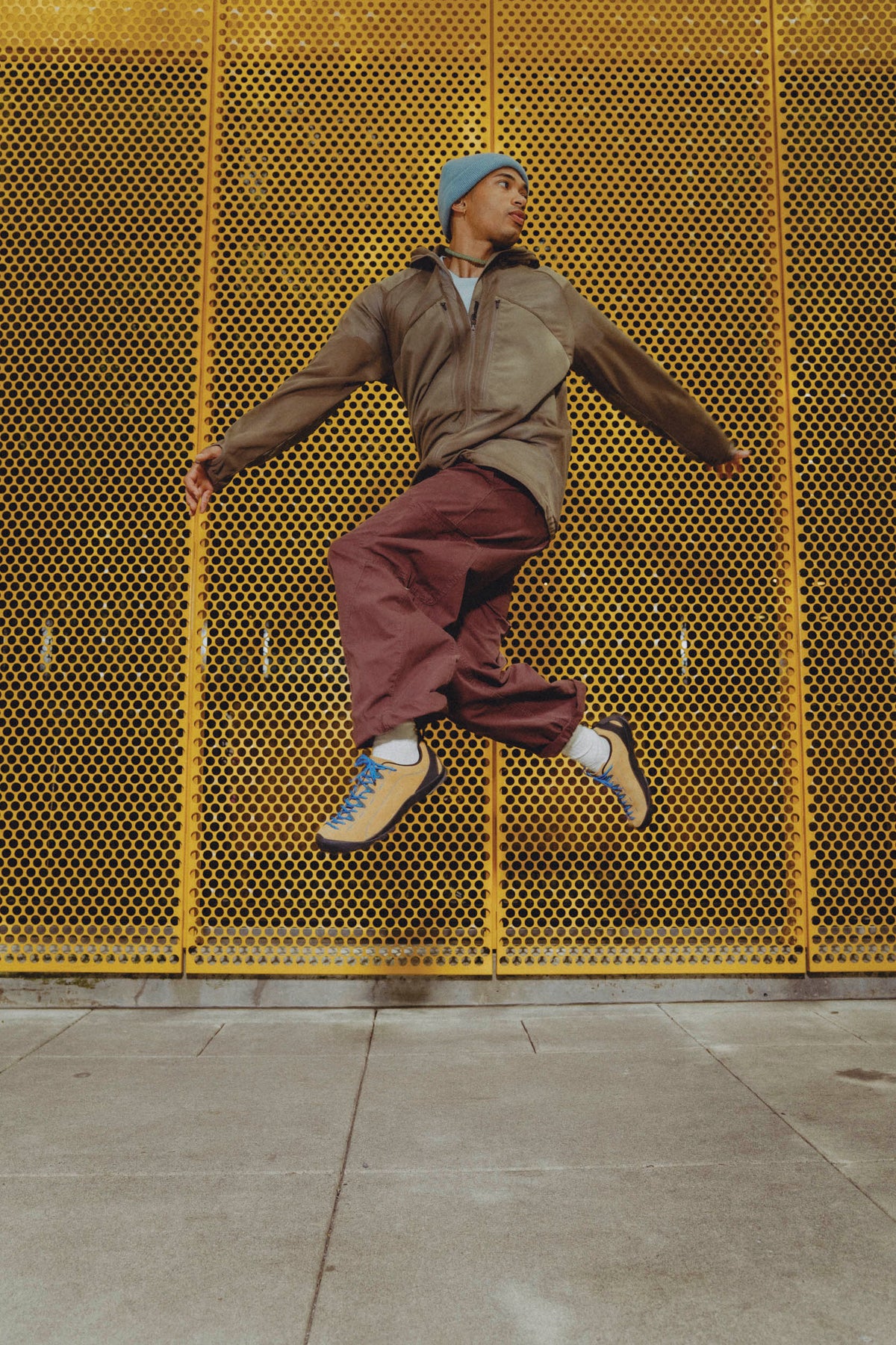 A person mid-jump in front of a bright yellow perforated metal wall, wearing a brown jacket