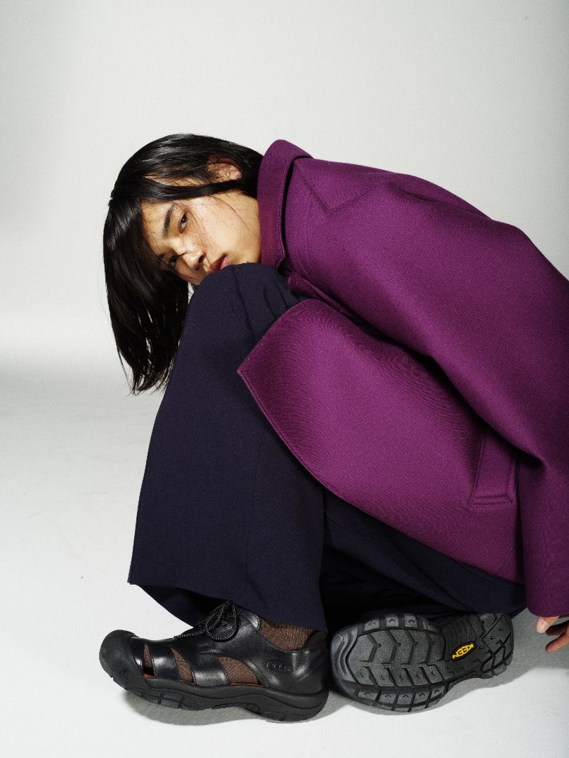 A model crouches on a light gray studio floor wearing an oversized deep purple coat, dark navy wide-leg trousers, and black Vibram sandals.