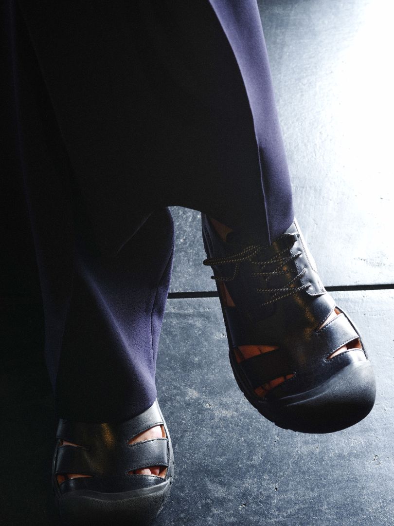 A close-up shot of a person's feet wearing black leather cutout sandals with orange lining, paired with dark trousers, standing on a dark floor.