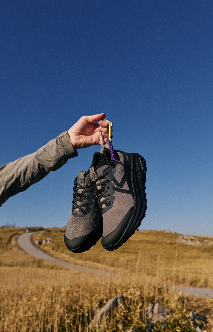 A person's hand holding a pair of keen grey and black trail running shoes.