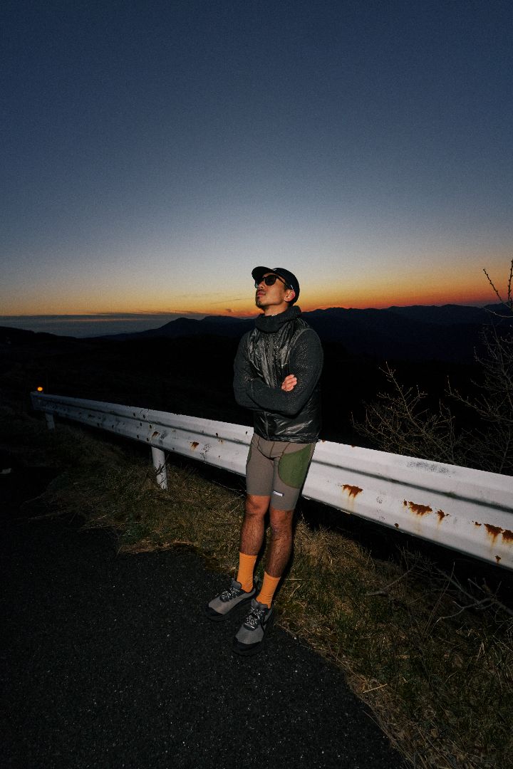 A man wearing a black jacket, green shorts, and keen trail running shoes looking up at the sky 