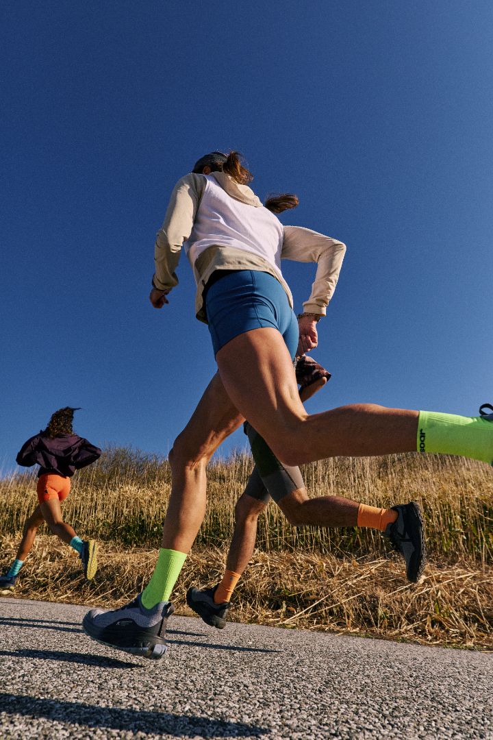 A low-angle action shot of a female trail runner from behind, wearing a white long-sleeve top, blue shorts, and bright neon green socks, running on a paved road with dry grass and a clear blue sky.
