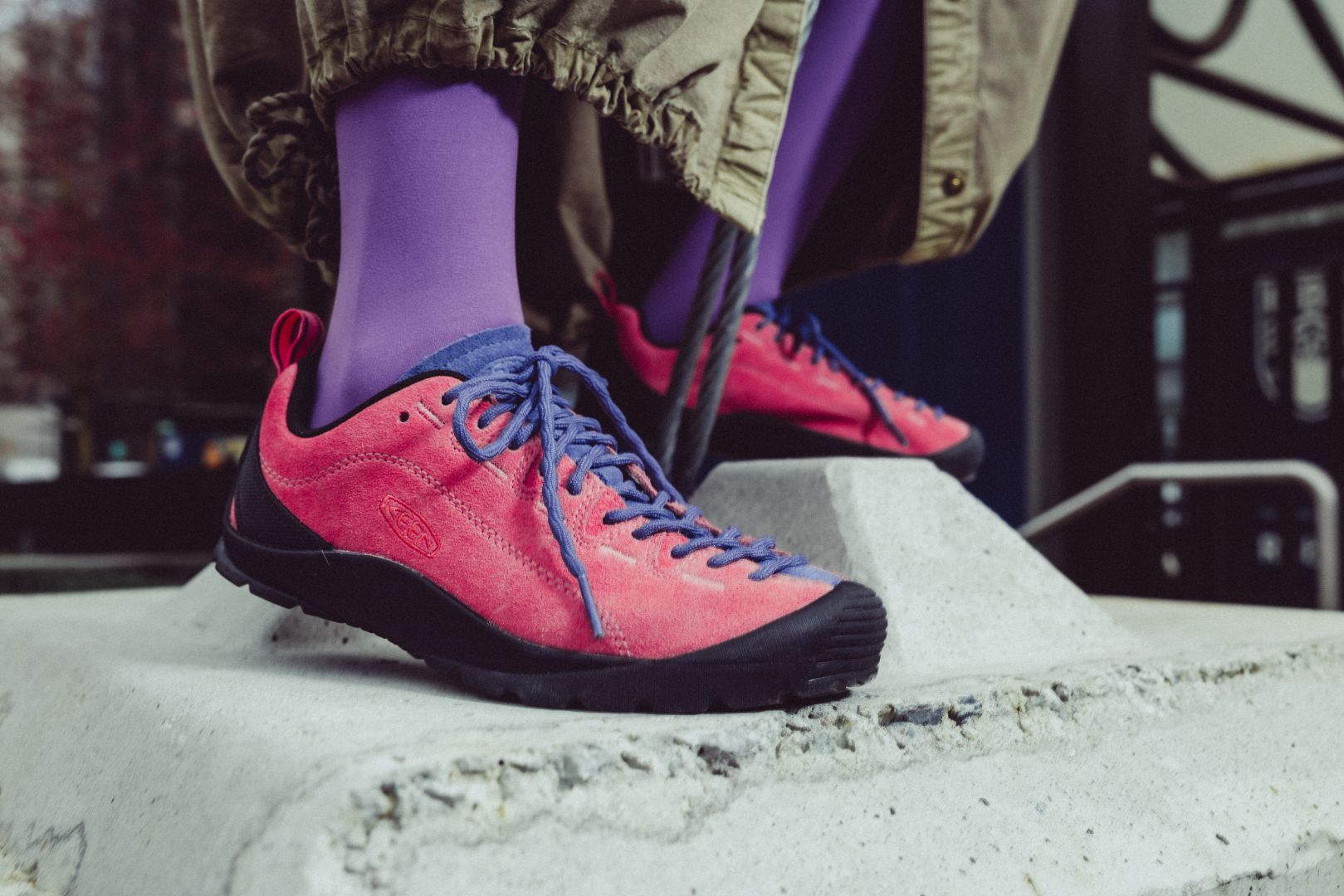 A close-up of a person wearing bright pink-red suede sneakers with blue laces and purple socks, standing on a concrete ledge outdoors.