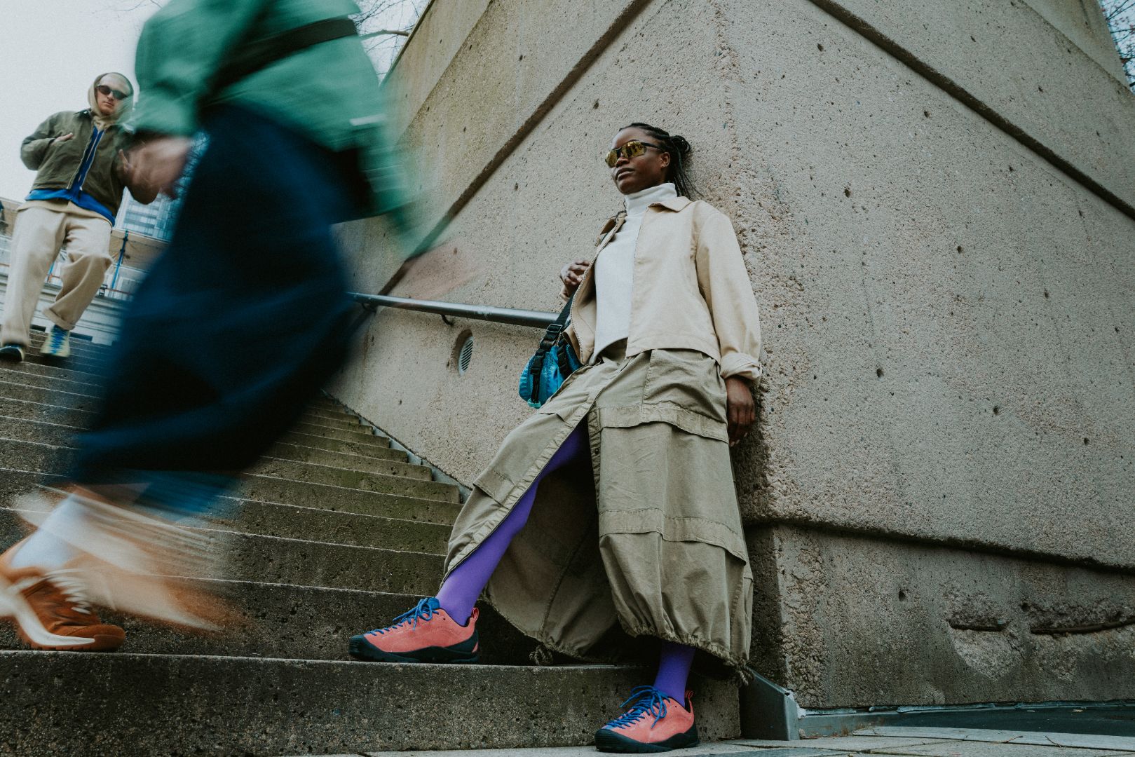 A woman in a beige jacket, oversized cargo pants, bright purple socks, and pink sneakers. Another person is blurred in motion in the foreground