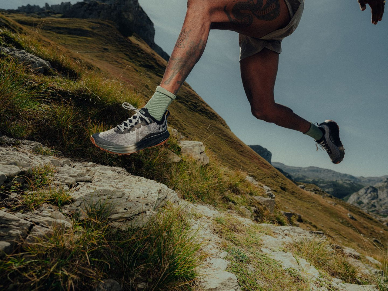 Person trailing running on a mountain trail with rocky terrain and grassy slopes. They are wearing Keen Seek trail running shoes in a black/grey colour.