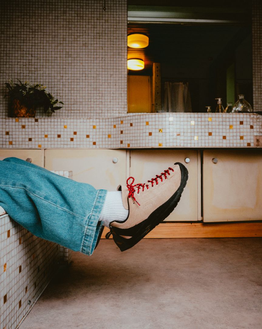 Person wearing Keen Jasper sneakers with red laces and blue jeans, lying in a tiled bathroom.