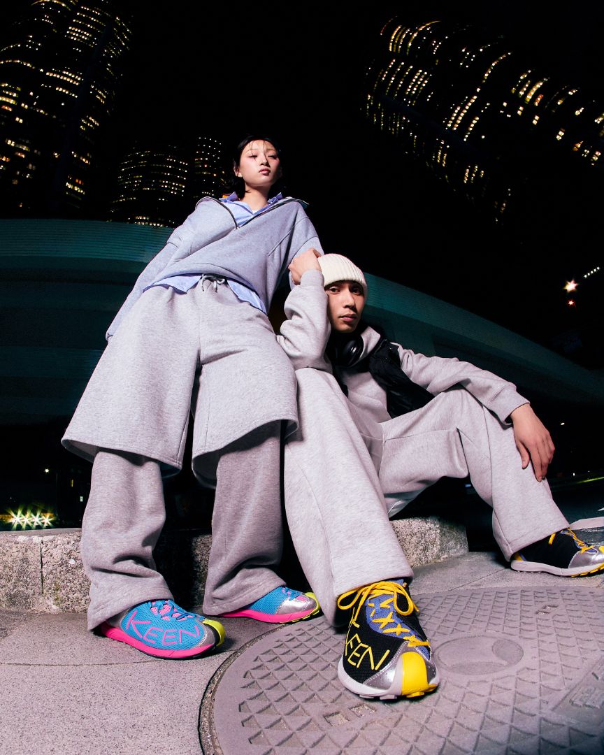 Two models in matching oversized light gray tracksuits pose together at night on a city ledge, with illuminated skyscrapers in the background. One model wears bright multicolor sneakers in blue, pink, and yellow, while the other wears black and yellow sneakers.