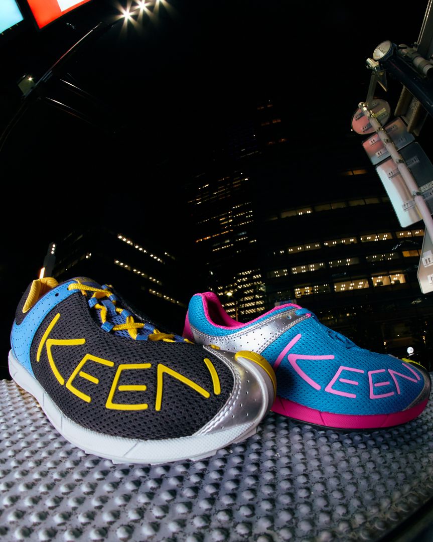 Two KEEN brand athletic shoes displayed on a textured surface at night with a city skyline in the background. One shoe is black with yellow lettering and blue laces, and the other is blue with pink lettering and a metallic silver toe cap.