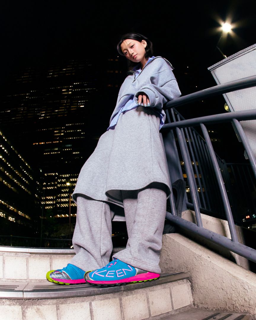 A person leaning on an outdoor night-time staircase railing, wearing a light blue oversized hoodie, a long gray coat, matching gray sweatpants, and bright blue sneakers with pink soles and 'KEEN' branding.