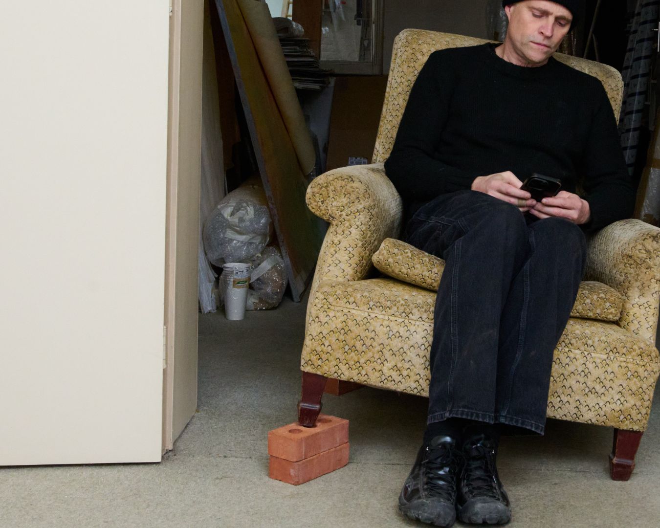 Man sitting in a patterned armchair using a smartphone in a cluttered room. The chair is balanced on a couple of bricks. The man is wearing the black Phileo x Keen collab  shoes 