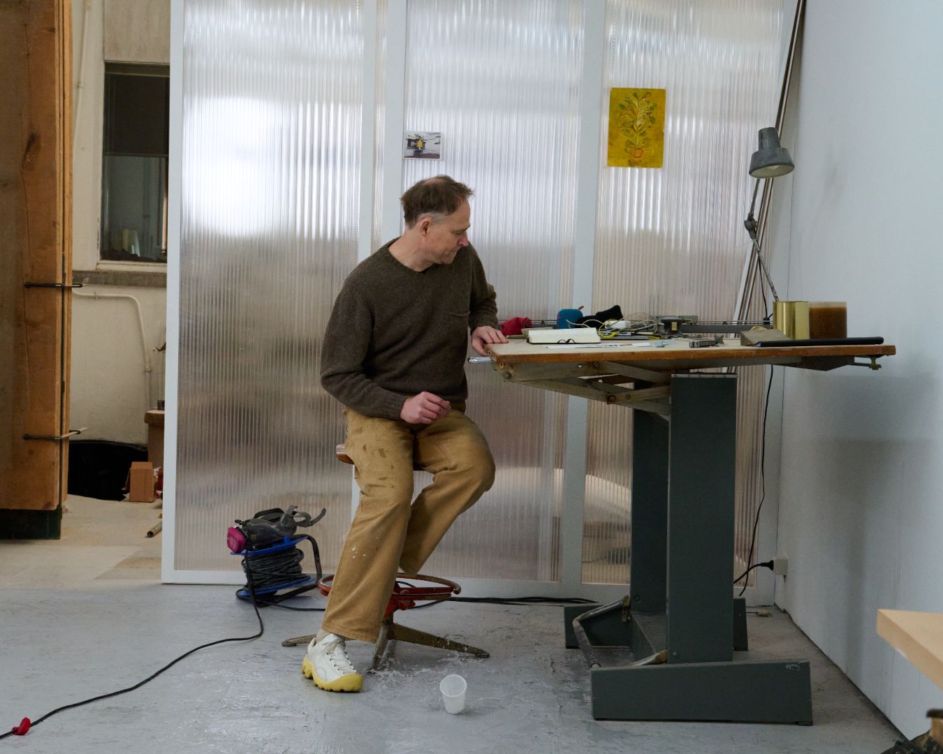 Man at his desk in a workshop he is wearing a brown jumper and tan coloured pants. On his feet he is wearing White Phileo X Keen Collab shoes