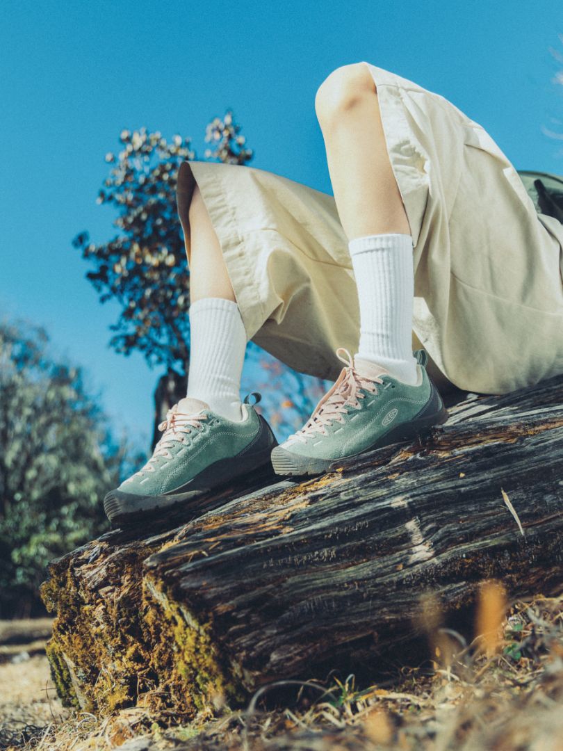 Person wearing light green Jasper sneakers and white socks sitting on a log outdoors with trees in the background.