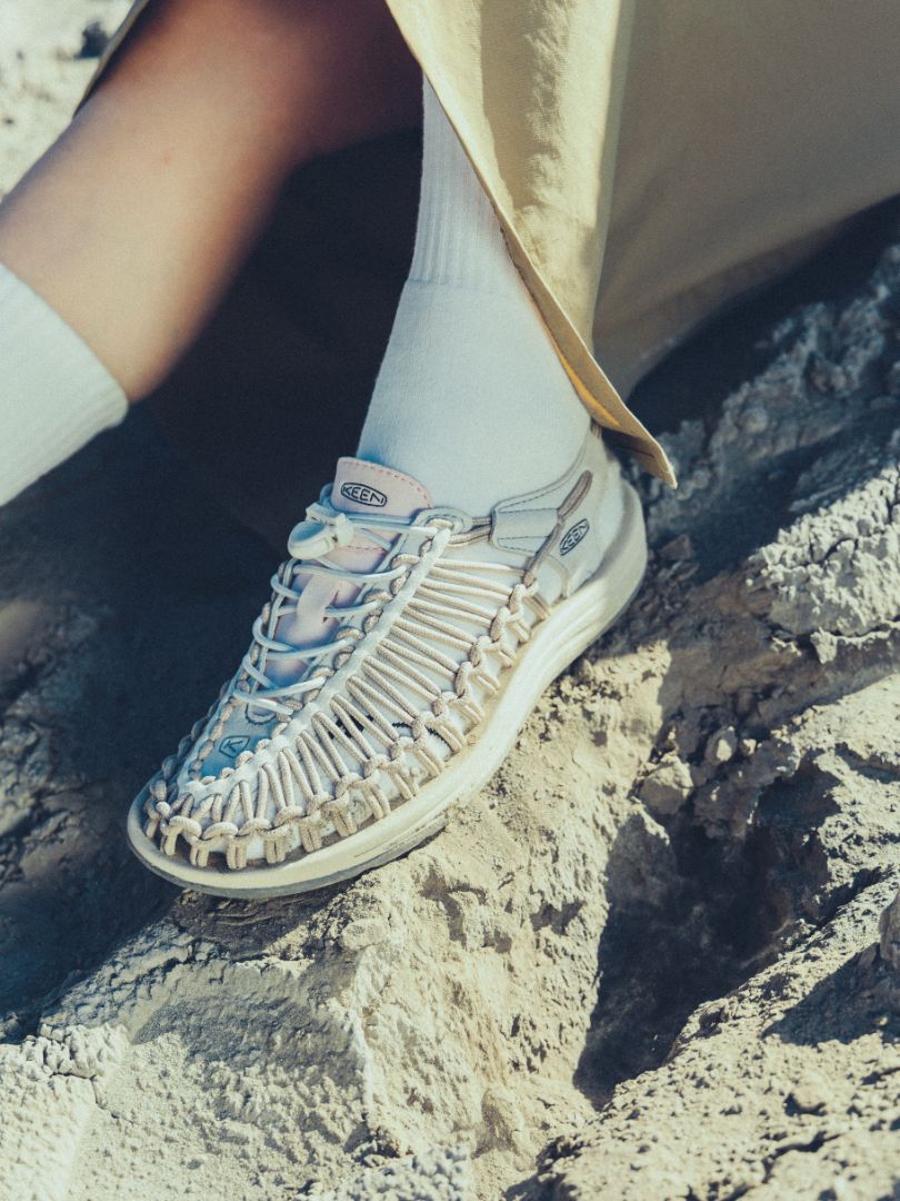 Close‑up of KEEN UNEEK footwear highlighting the signature interwoven cord design, secure lace system, textured outsole, and light‑colored rubber sole on rocky ground.