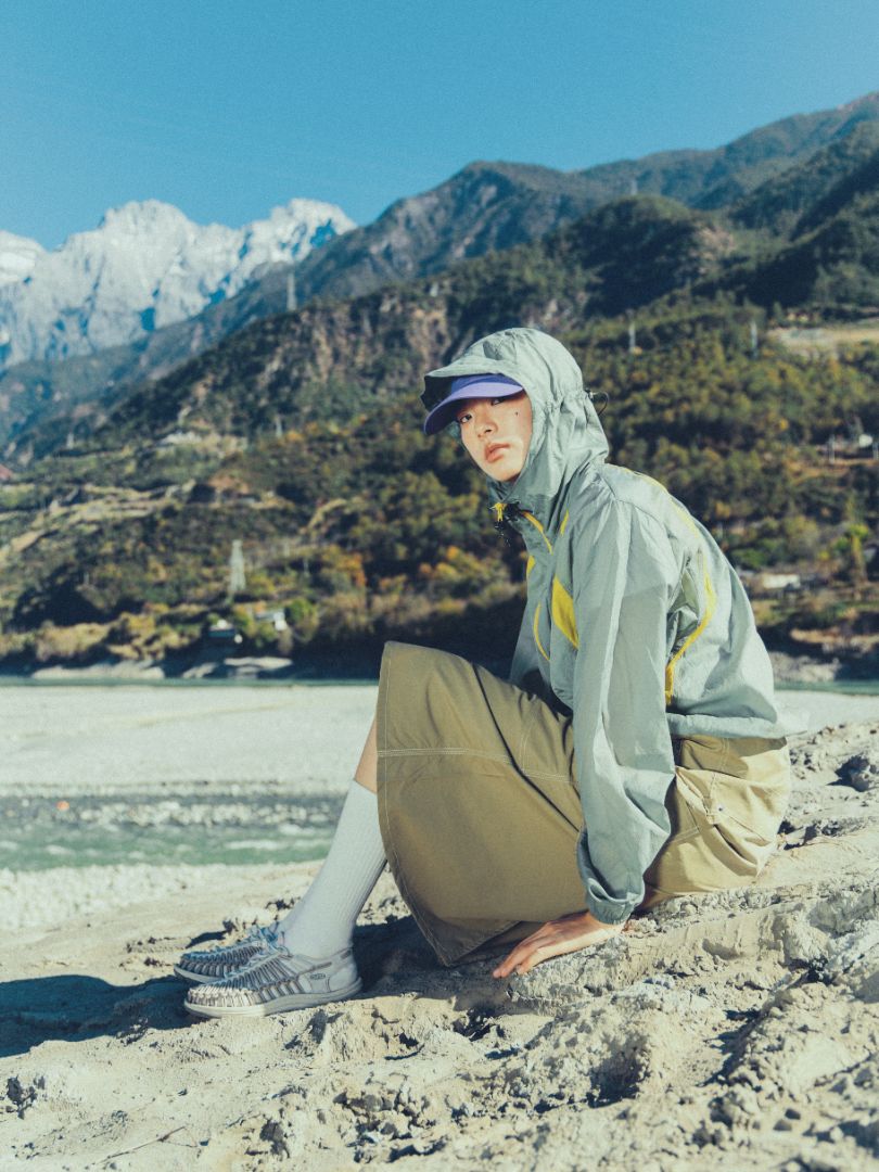 KEEN UNEEK footwear visible from the side while seated on sand, emphasizing the foot‑hugging cord upper, supportive sole, and versatility in an outdoor mountain setting