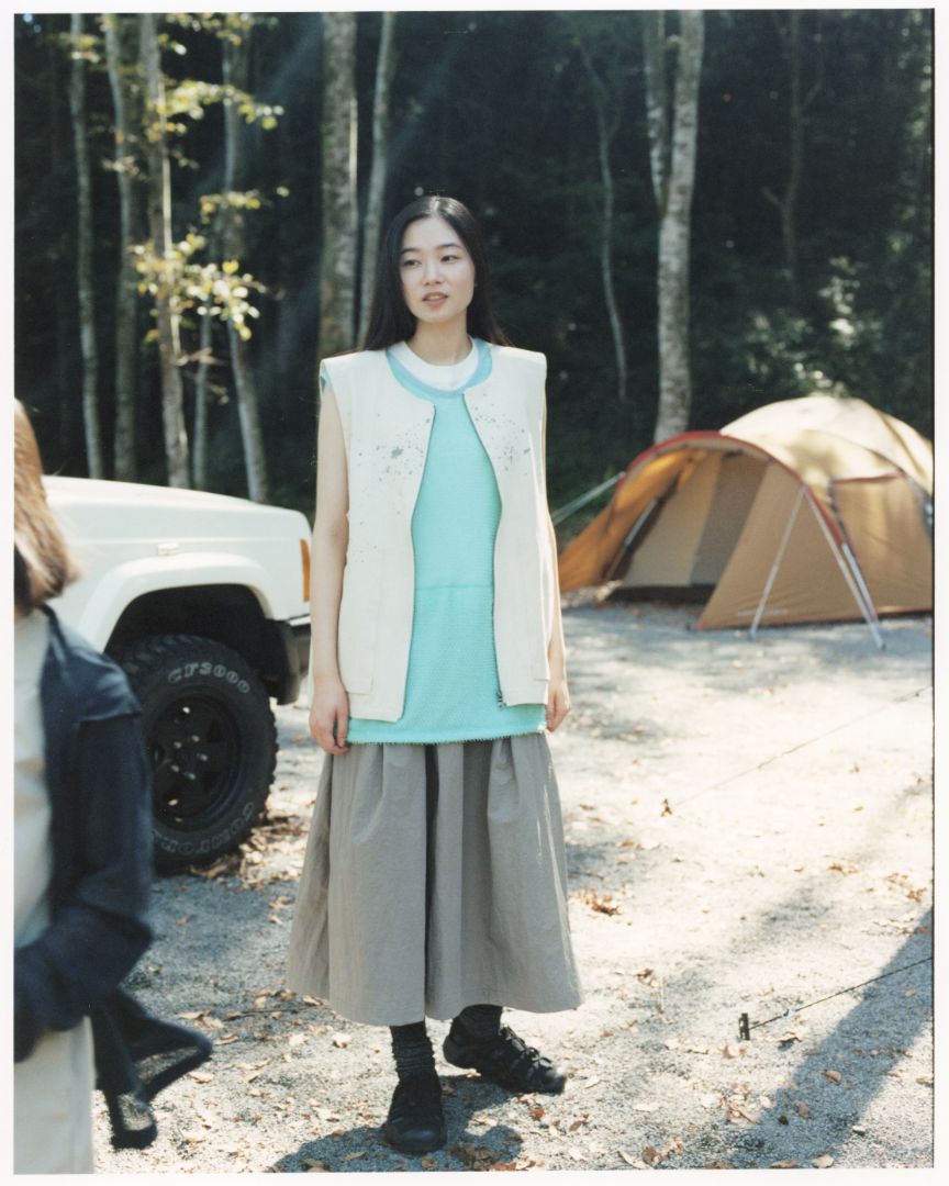 Person wearing black keen x snow peak hyperport sandals outdoor sandals standing at a forest campsite with tents and a parked vehicle in the background.