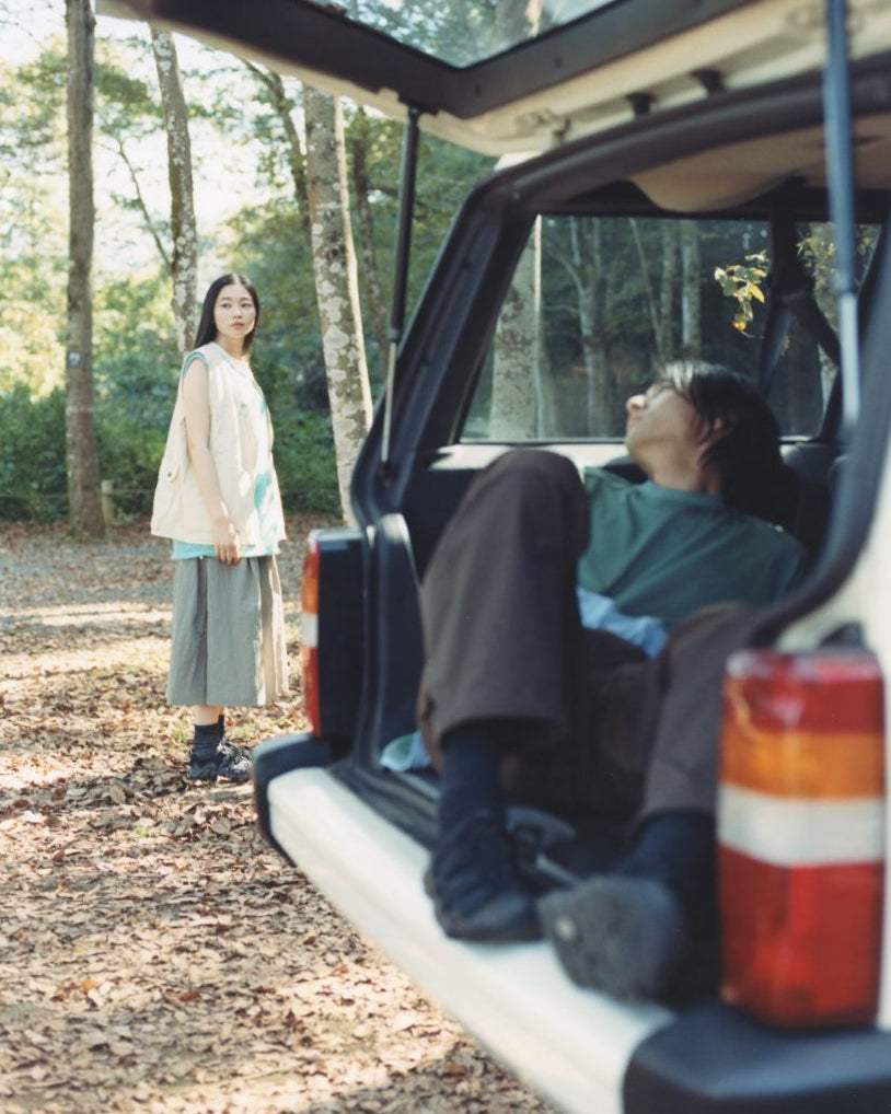 Two people in a forest setting, one sitting in an open car trunk and the other standing nearby.