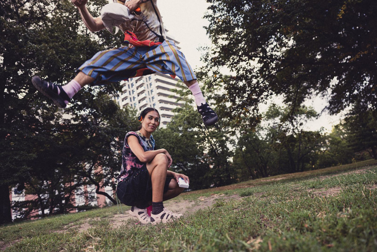 Person in mid-air performing a trick wearing Keen Newport Sandals in black with another person sitting on the ground in a park setting wearing Newport Sandals in a Beige colour