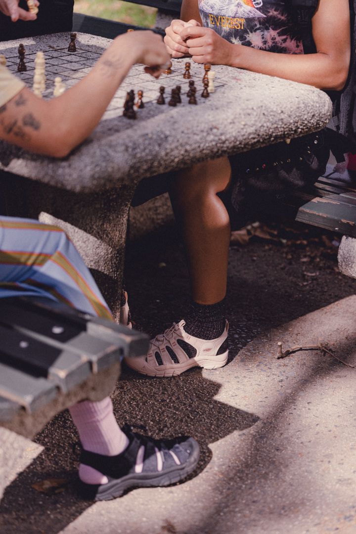 Two people sat playing chess outdoors on a bench. One person is wearing Newport Sandals in Black, the other is wearing Newport Sandals in Beige.
