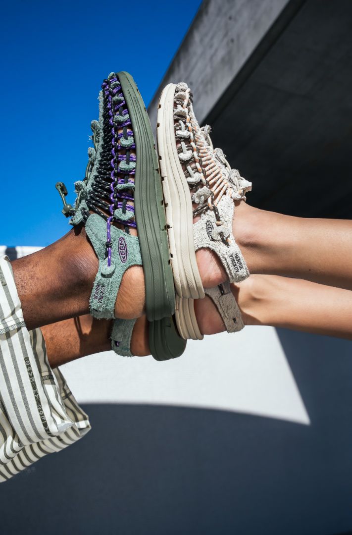 Two pairs of feet wearing rugged KEEN UNEEK x Henrik Vibskov sandals—one green and one beige—pressed together sole to sole, photographed outdoors against a blue sky and concrete building