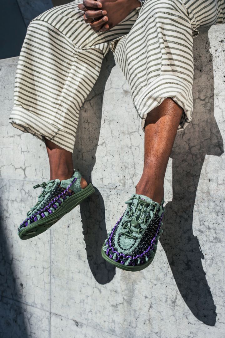 Person sat on a wall wearing the collab spring green Henrik Vibskov X UNEEK sneakers with purple accents on a concrete surface