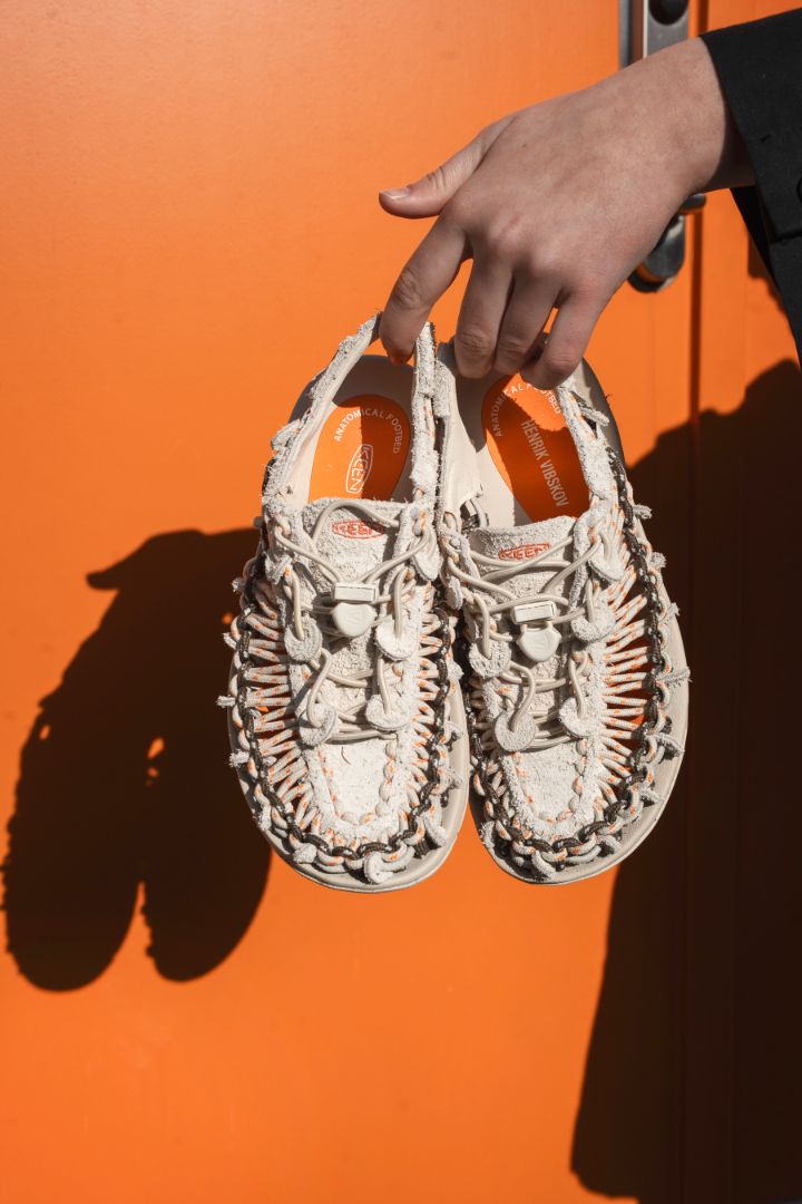 Henrik Vibskov x KEEN UNEEK sandals held by the heels against a bright orange background, highlighting the signature braided cord upper, secure lace system, and collaborative design details.