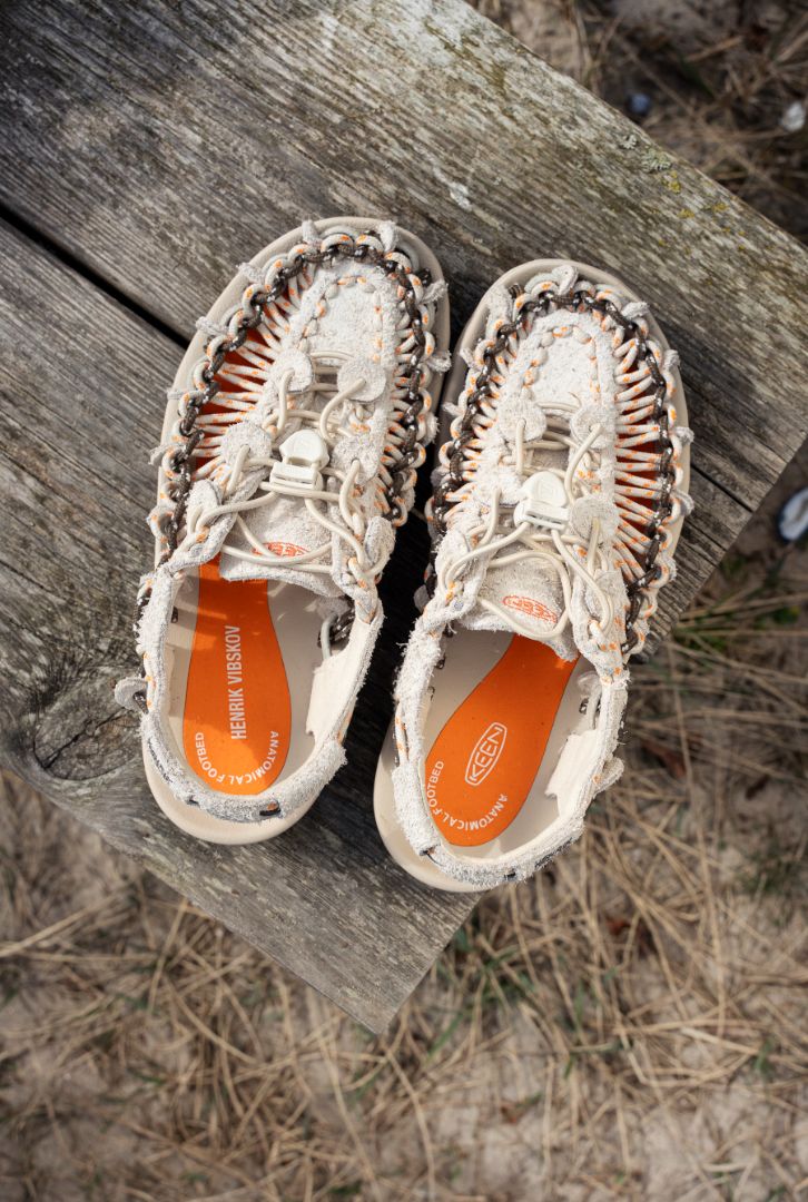 Top‑down view of Henrik Vibskov x KEEN UNEEK sandals on a weathered wooden surface, emphasizing the interwoven cord construction, cushioned footbeds, and bold orange collaborative insoles