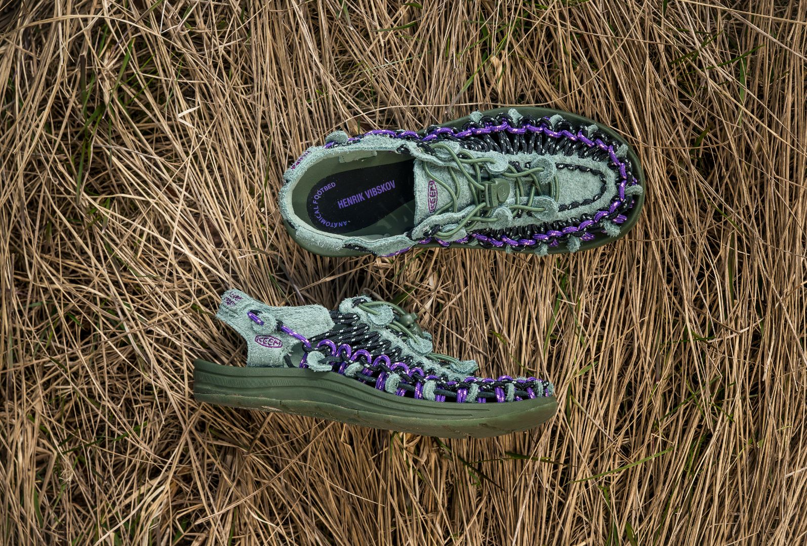 Henrik Vibskov x KEEN UNEEK sandals arranged on dry grass, showcasing the suede‑like upper, green and purple braided cord accents, and elevated sole design from the collaboration