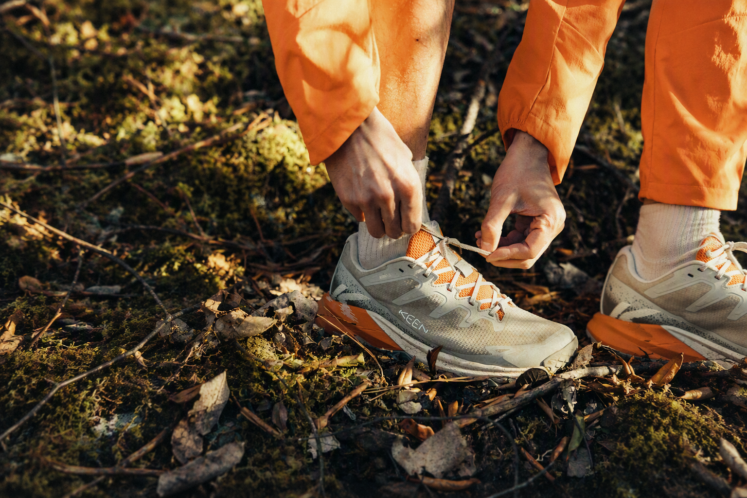 A close-up outdoor shot of a person tying the laces of a light gray and orange running shoe while wearing orange pants on a mossy forest floor.