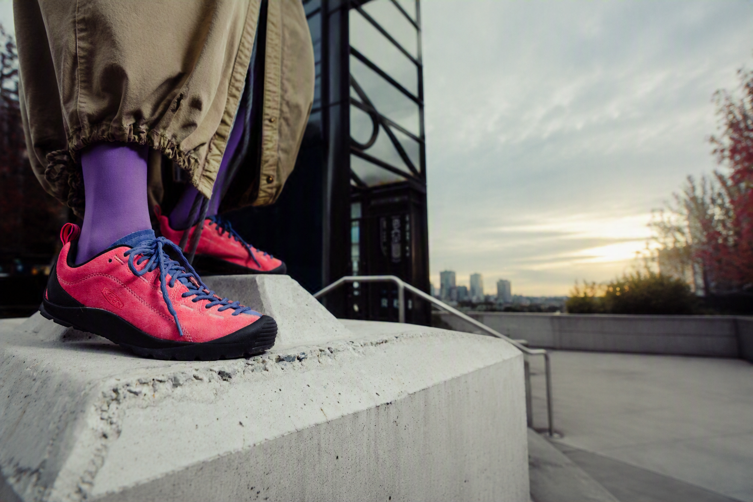 A close-up lifestyle shot of a person's legs and feet wearing bright pink sneakers with blue laces, purple socks, and khaki pants, standing on a concrete ledge in an outdoor urban setting.
