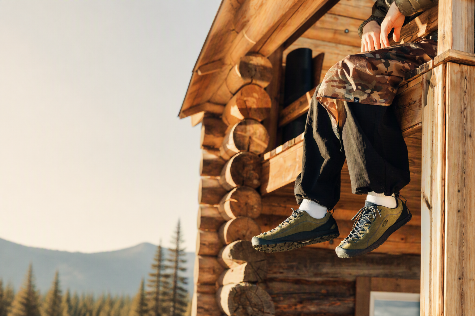 A person sitting on the wooden porch of a log cabin wearing olive green hiking shoes, black pants, and camouflage top, with a mountain landscape in the background.