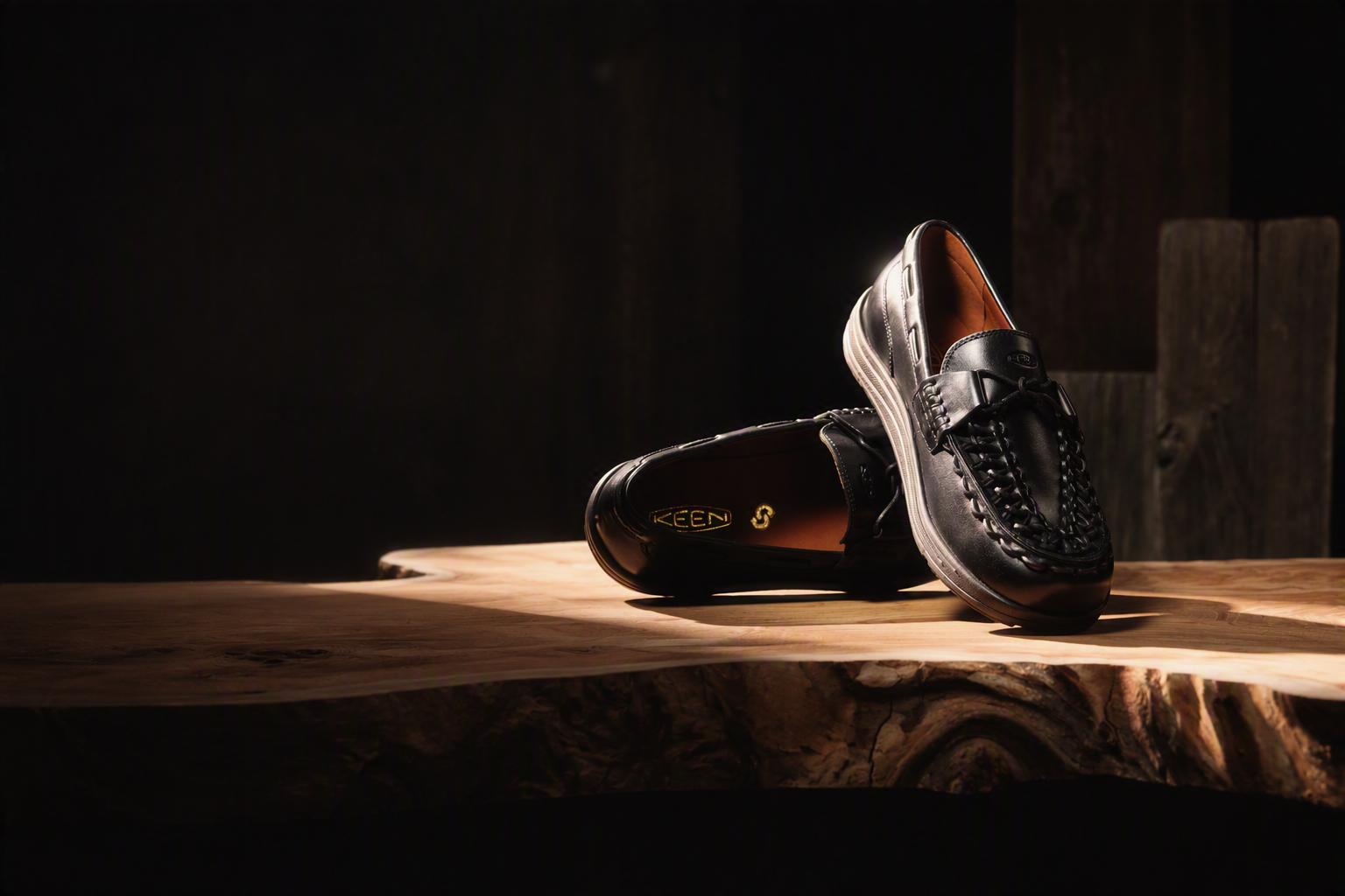 A pair of black leather loafers with braided detailing, displayed on a rustic wooden slab against a dark background. One loafer is standing upright while the other lies on its side, showing the brand name 'KEECH' on the insole.