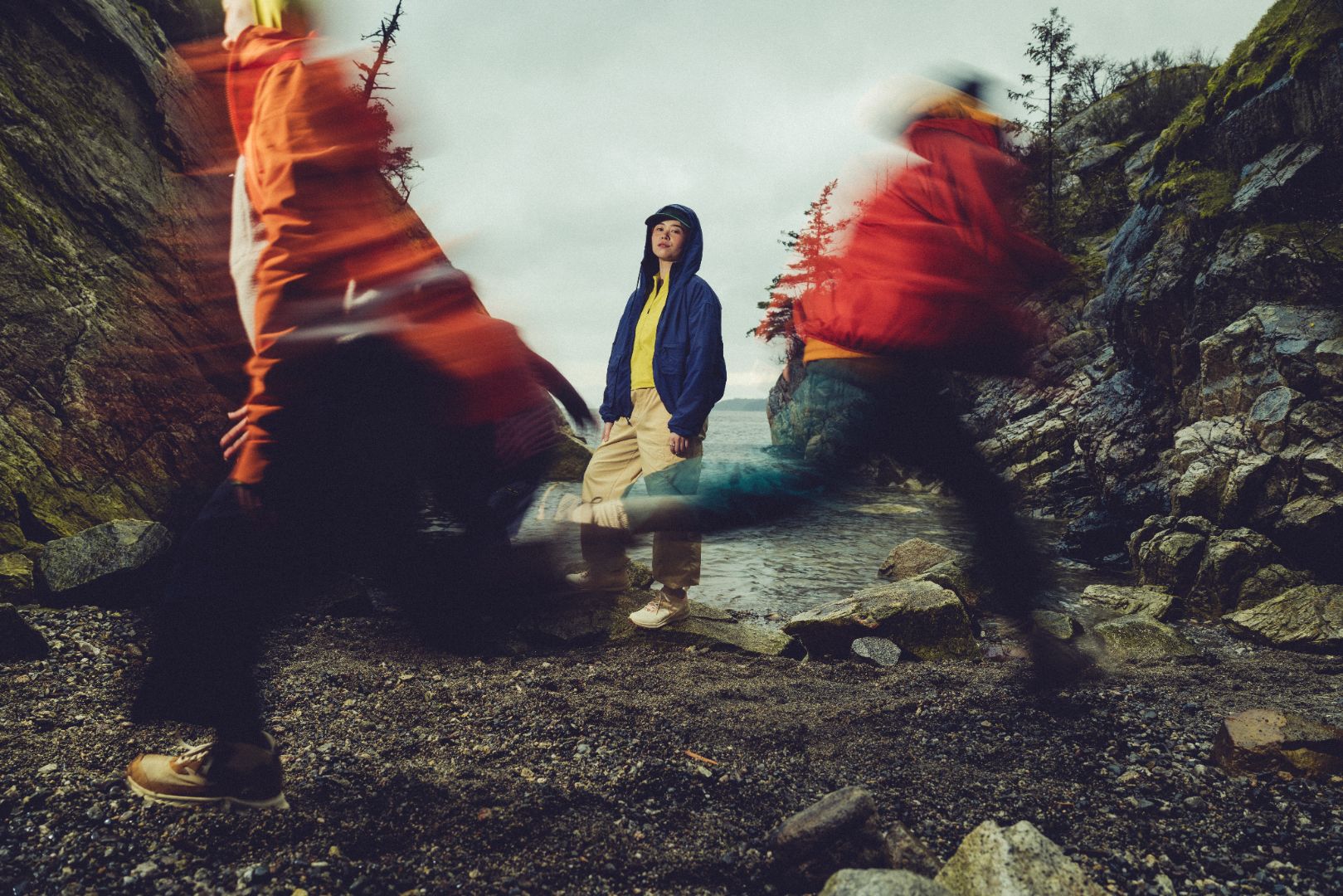Three people in colorful jackets standing on a rocky path with a mountainous background.