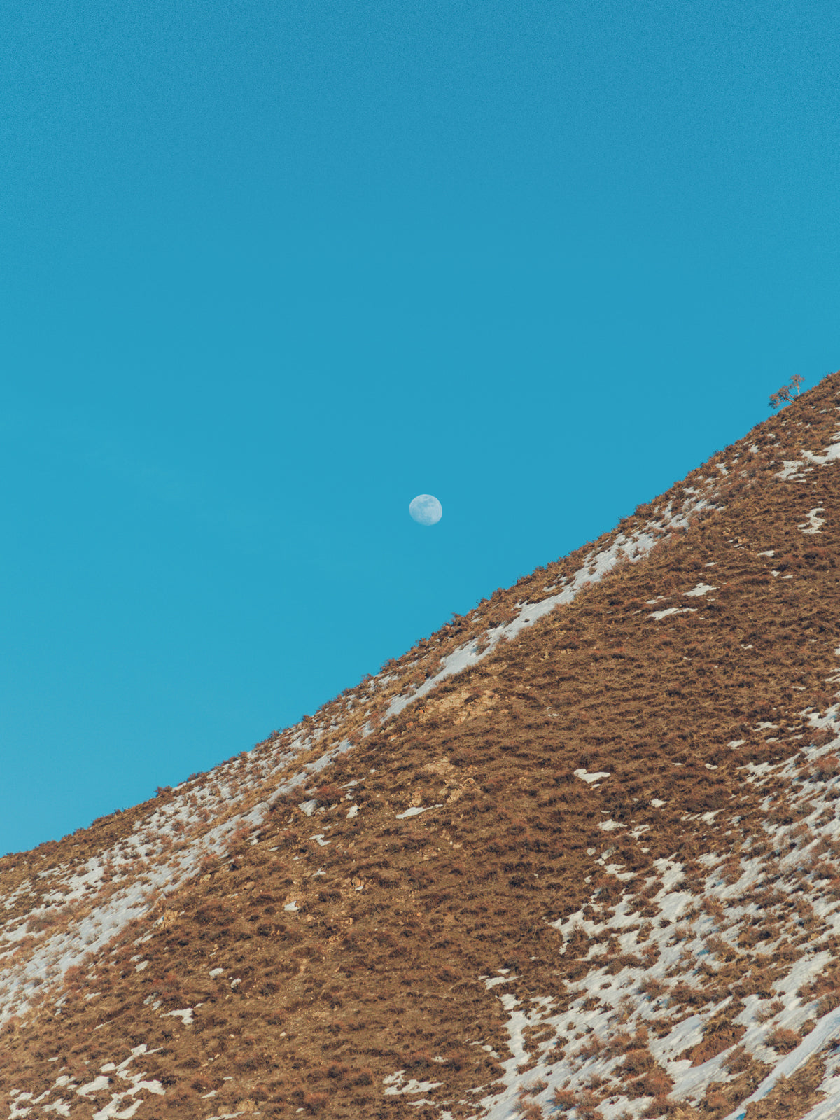 A vast blue sky with a small moon and tan mountainside 