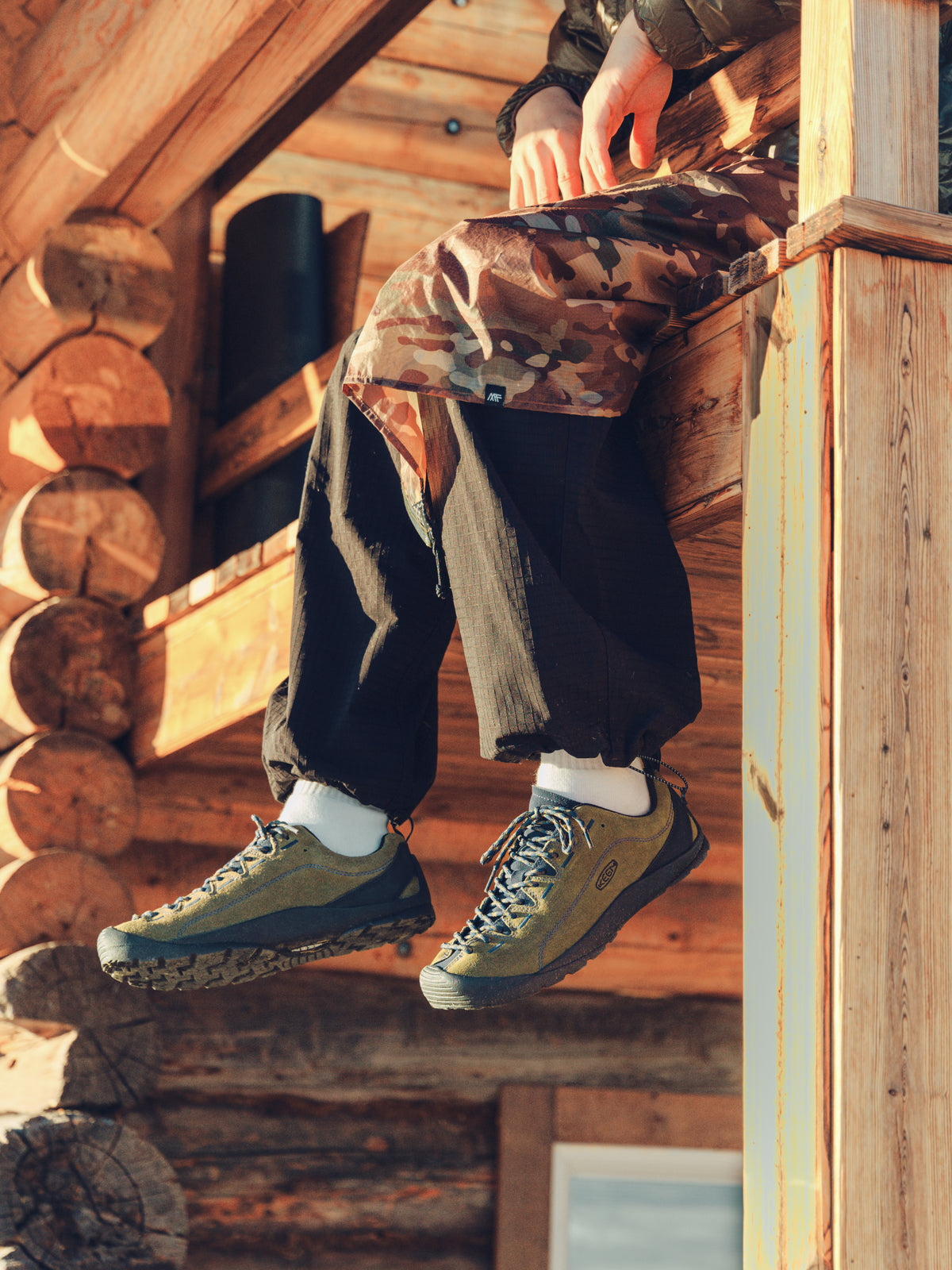 A person sitting on a rustic wooden log cabin porch, wearing olive green and blue hiking shoes