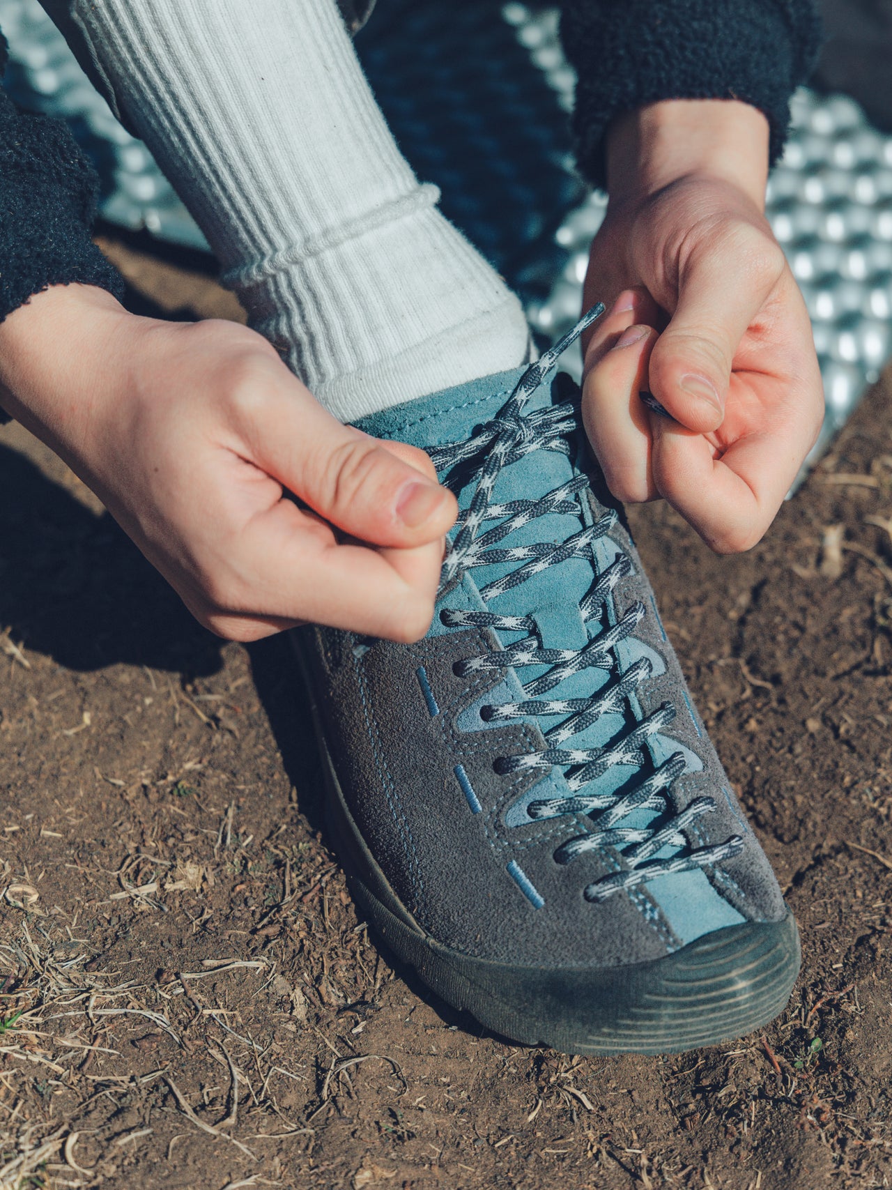 A close-up of a person's hands tying the laces of a blue jasper suede hiking shoe, with a white ribbed sock and dirt ground visible.
