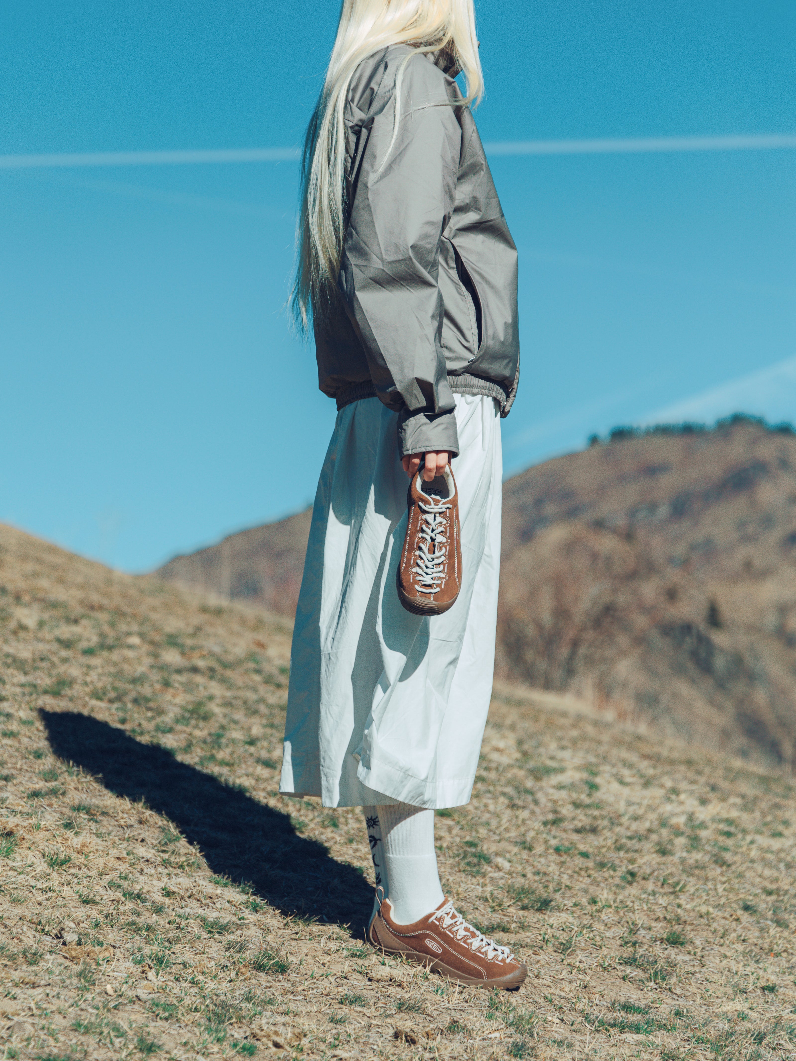 A person standing outdoors on a grassy hill under a clear blue sky, wearing a gray jacket, a long light-colored skirt, white socks, and brown lace-up shoes, while holding a brown shoe in one hand.