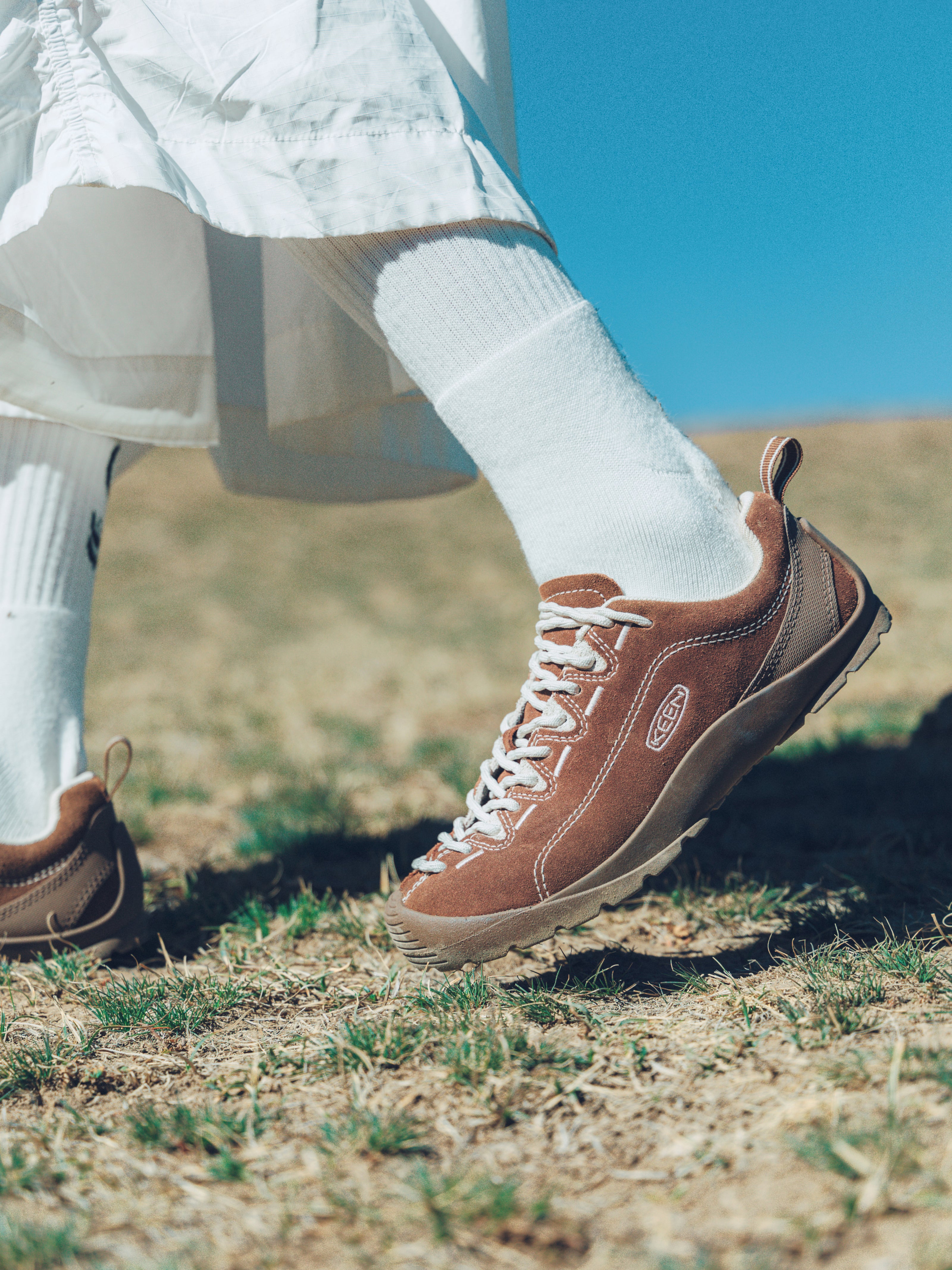A person outdoors wearing brown suede K-Swiss sneakers with white socks, standing on dry grass under a clear blue sky.
